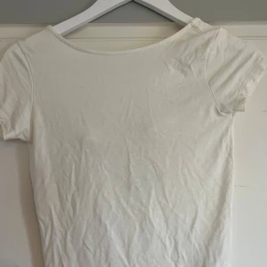 Vit topp med rosett från 157 - Säljer en söt vit topp från 157 i storlek XS. Toppen har korta ärmar och en djup rygg med en fin rosettdetalj. Perfekt för varma dagar och enkel att matcha med olika outfits. Den är lite genomskinlig