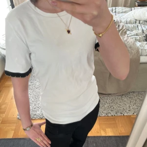 Vit t-shirt med svarta volangdetaljer - Säljer nu den slutsålda Zara t-shiten med unika svarta detaljer och guldfärgade inslag på ärmsluten. Perfekt för dig som vill ha något enkelt men ändå med en twist. Den är i storlek M men passar mig som vanligtvis har xs och s. Säljer då den inte kommit till användning, ändast använd 1 gång. Kontakta mig om frågor!💞💓 (Kolla med mig först då jag är i min stuga och inte vet om jag kommer hinna posta just denna) 💓