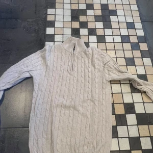 Beige stickad tröja med dragkedja från John Henric - Säljer en beige stickad tröja från John Henric med halv dragkedja vid halsen. Tröjan har kabelstickat mönster och ribbade muddar vid ärmslut och nederkant. Perfekt för sommarkvällarna som lager ovanför tex en skjorta, storlek S