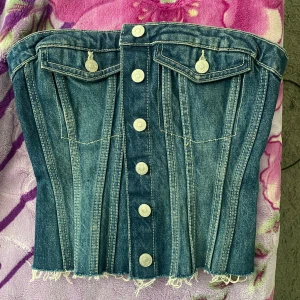 Jeans korsettopp med knappar - Säljer en snygg jeans korsettopp med silverfärgade knappar framtill och två dekorativa fickor. Toppen har markerade sömmar och rå kant nertill för en cool look. Den är bara testad hemma men pris lappen är av. Så den är som ny!! Storlek. S men passar xs också