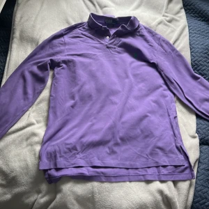 Lila långärmad pikétröja från Polo Ralph Lauren - Säljer en lila långärmad pikétröja från Polo Ralph Lauren med klassisk krage och knappar framtill. Tröjan har en broderad grön logga på bröstet och är tillverkad i bomull. Perfekt för en stilren och avslappnad look.