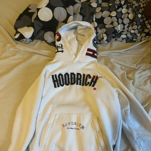 Vit hoodie från Hoodrich - Säljer en vit hoodie från Hoodrich med svart och röd logga på bröstet och detaljer på huvan. Stor magficka framtill och tryck även på ryggen. Perfekt för dig som gillar streetwear och vill sticka ut med snygga detaljer.