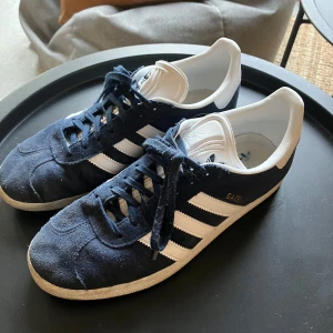 Adidas Gazelle mörkblå sneakers - Säljer ett par klassiska Adidas Gazelle sneakers i mörkblå mocka med vita detaljer och vita ränder på sidorna. 