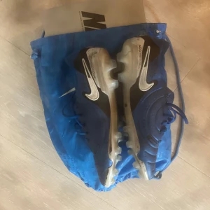 Blåa Nike fotbollsskor med vit swoosh - Säljer ett par blåa Nike fotbollsskor med vit swoosh och platt sula med dobbar. Skorna har snörning och är perfekta för dig som vill sticka ut på planen. Levereras med blå Nike påse 