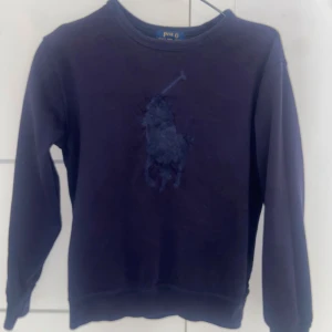 Mörkblå sweatshirt från Polo Ralph Lauren - Mörkblå sweatshirt med rund hals från Polo Ralph Lauren. Tröjan har ett stort broderat logomärke på bröstet i samma färg som tyget. Klassisk och enkel design med långa ärmar och mjukt material.