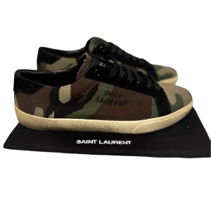 Saint Laurent skor  - Säljer ett par Saint Laurent sneakers med camouflage-mönster och svarta detaljer. Skorna har snörning, rund tå och platt sula. Perfekta för dig som vill ha en cool och stilren look med streetkänsla.