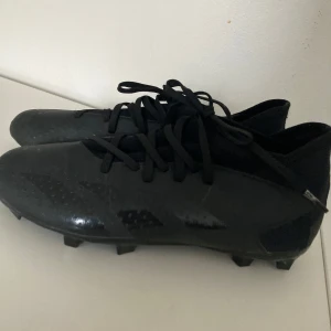 Svarta Adidas Predator fotbollsskor - Säljer ett par svarta Adidas Predator fotbollsskor med snörning. Skorna har en låg profil och är tillverkade i syntetmaterial med mönstrad ovandel för extra grepp. Perfekta för spel på gräsplaner. Köpt för 1100 