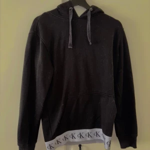 Svart hoodie från Calvin Klein Jeans - Svart hoodie från Calvin Klein Jeans med huva och dragsko. Nederkanten har en vit detalj med CK-logga runt om. Perfekt för en chill och stilren look.