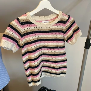 Randig stickad topp från Vero Moda - Superfin stickad topp från Vero Moda med korta ärmar. Tröjan har ränder i beige, rosa, svart och vitt samt virkade detaljer och vågig kant vid ärmslut och nederkant. Perfekt för dig som gillar färg och mönster!💞