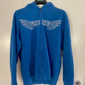 Blå hoodie med strasstenar - Säljer en blå hoodie med glittriga strasstenar i form av vingar. Tröjan har dragkedja framtill och huva med snörning. Perfekt för dig som vill sticka ut!
