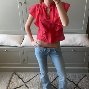 Röd volangblus med kort ärm - Säljer en röd blus med volangdetaljer och korta ärmar. Blusen har knappar framtill och är croppad i modellen. Perfekt för dig som vill ha en färgstark och luftig topp till sommaren.
