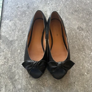 Svarta ballerinaskor med rosett - Säljer ett par svarta ballerinaskor med rund tå och dekorativ rosett framtill. Skorna har öppen tå och platt sula. Så söta och sköna!!