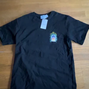 Svart t-shirt från Casablanca med tryck - Svart t-shirt från Casablanca med färgglatt tryck av en byggnad och gröna blad på bröstet och ryggen. T-shirten har rund hals och korta ärmar. Perfekt för dig som gillar unika prints och streetwear.