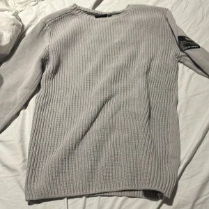 Ljusgrå stickad tröja från Sail Racing - Säljer en ljusgrå stickad tröja från Sail Racing Antarctica-kollektionen. Tröjan har rund halsringning, ribbade muddar och diskreta logodetaljer på ärmen och nacken. Perfekt för kyligare dagar.