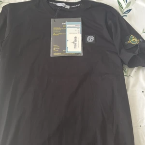 Svart t-shirt från Stone Island - Säljer en svart t-shirt från Stone Island med klassisk logotyp på bröstet och ärmen. T-shirten har rund hals och korta ärmar. Perfekt för dig som gillar streetwear och vill ha något stilrent med ikonisk branding.
