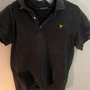 Svart pikétröja från Lyle & Scott - Säljer en svart pikétröja från Lyle & Scott med klassisk krage och knappar framtill. Tröjan har korta ärmar och den ikoniska gula fågel-loggan broderad på bröstet. Perfekt för en stilren och avslappnad look.