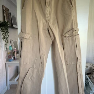 Beiga cargobyxor från Pull&Bear - Säljer ett par beiga cargobyxor från Pull&Bear. Byxorna har flera fickor på sidorna och en rak passform. Perfekta för en avslappnad och trendig stil. Modellen är Tall och passar dig som vill ha lite extra längd.