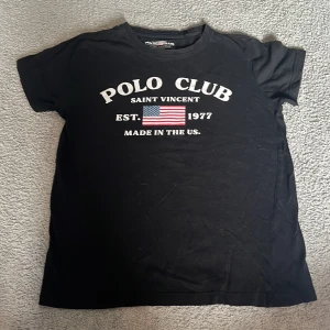 Svart t-shirt från Polo Club - Svart t-shirt från Polo Club med vit text och amerikansk flagga tryckt på bröstet. Klassisk rund hals och korta ärmar. Perfekt för en avslappnad och sportig stil.