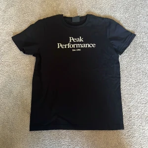 Svart t-shirt från Peak Performance - Svart t-shirt från Peak Performance med vit logotyp och text 'Est. 1986' på bröstet. Klassisk rund halsringning och korta ärmar. Perfekt för en clean och sportig stil.