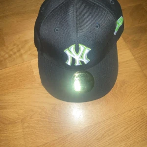 Svart New Era keps med limegrön NY-logga - Säljer en svart keps från New Era med broderad New York Yankees-logga i vitt och limegrönt framtill. Kepsen har platt skärm och broderad New Era-logga på sidan. MLB-logga baktill. Perfekt för dig som gillar sportig stil.