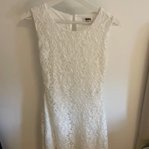 Vit spetsklänning från DM - Säljer en elegant vit klänning från Dennis Maglic i storlek S. Klänningen är ärmlös och har ett vackert blommigt spetsmönster samt rund halsringning. Perfekt för somriga tillfällen eller studentfirande. Använd en gång.