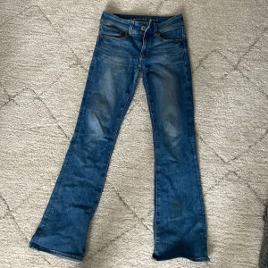 Blå bootcut jeans från American Eagle - Säljer ett par klassiska blå bootcut jeans från American Eagle med Next Level Stretch. Jeansen har fem fickor, knapp och dragkedja framtill samt snygga slitningar för en avslappnad look. Med mönstrade fickor 