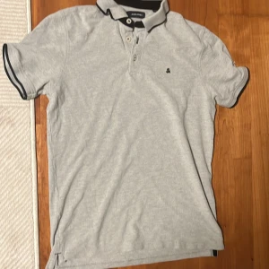 Ljusgrå piké t-shirt från Jack & Jones - Snygg ljusgrå piké t-shirt från Jack & Jones med svart kant på krage och ärmslut. Klassisk knappslå med tre knappar och liten broderad logga på bröstet. Perfekt för en stilren och avslappnad look.