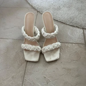 Vita klackade sandaler med flätade remmar - Säljer ett par trendiga vita sandaler med bred klack och två flätade remmar över foten. Skorna har fyrkantig tå och öppen häl, perfekta för att lyfta din outfit.