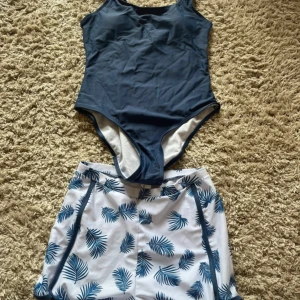 Blå baddräkt och vita badshorts med blått blad-mönster - Säljer en mörkblå enkel baddräkt med smala axelband och ett par vita badshorts med blå palmbladsmönster. Shortsen har blå kantband och är höga i modellen. Perfekt för strand eller pool!