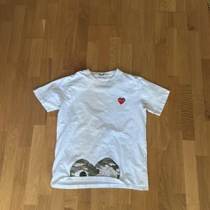 Vit t-shirt från Comme des Garçons Play med camo-detalj - Vit t-shirt från Comme des Garçons Play med rött hjärta på bröstet och camouflagemönstrad detalj längst ner. Klassisk passform och korta ärmar. Perfekt för dig som gillar streetwear och unika detaljer.
