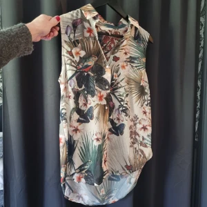 Blommig ärmlös skjorta från H&M - Säljer en tunn och luftig ärmlös skjorta från H&M i storlek 38. Skjortan har ett tropiskt blommönster med fjärilar och fåglar i grönt, beige och inslag av orange. Perfekt för varma dagar och enkel att styla med jeans eller shorts. Pris 50kr
