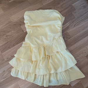 Ljusgul volangklänning - Supersöt ljusgul klänning med flera volanglager och axelbandslös design. Perfekt för somriga dagar eller speciella tillfällen. Klänningen har en kort längd och är tillverkad i ett lätt, flowy material som ger en luftig känsla.