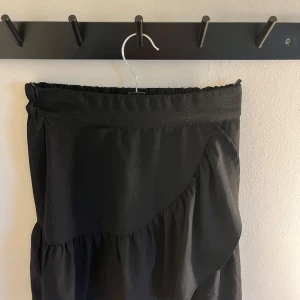 Svart kjol med volang - Snygg svart kjol med volangdetaljer från Lindex. Går en bit över knäna på mig som är 160. Elastisk midja och i storlek xs. Har däremot fått ett fåtal vita ”noppror” som syns på bilderna💕