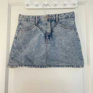 Blå jeanskjol från stradivarius  - Säljer en klassisk blå jeanskjol från stradivarius med femficksmodell och knappgylf. Kjolen är kort och har en rak passform, perfekt till sommaren. Snygga detaljer med synliga sömmar och bälteshällor.