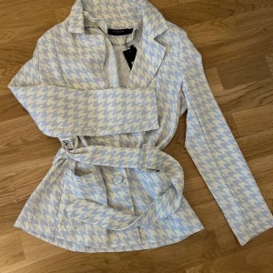 Ljusblå och vit rutig kavaj från In Vogue - Säljer en snygg kavaj i ljusblått och vitt hundtandsmönster från In Vogue. Jackan har lång ärm, knappar framtill och ett matchande bälte i midjan. Perfekt för dig som vill sticka ut med stilren design.