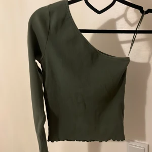 Grön ribbad oneshoulder topp -  grön croppad topp med ribbad struktur och oneshoulder-design. Toppen har lång ärm på ena sidan. ”Volang kant” vid midjan och ärmslutet.
