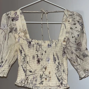 Blommig croppad blus med puffärmar -  croppad blus med blommigt mönster i lila och beige toner. Toppen har puffiga trekvartsärmar, smockad kropp och knytning i ryggen. 