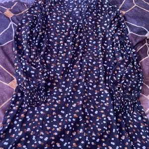 Mörkblå prickig klänning med långa ärmar - Säljer en mörkblå klänning med vita, ljuslila och rostbruna prickar. Klänningen har långa ärmar med smock vid ärmsluten och en lätt v-ringning. Materialet känns som polyester och klänningen har en lös passform.
