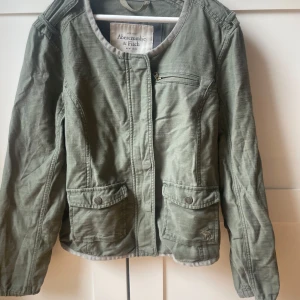 Grön jacka från Abercrombie & Fitch - Snygg militärgrön jacka från Abercrombie & Fitch med dragkedja och knappar framtill. Jackan har två stora fickor med lock och en bröstficka med dragkedja. Perfekt till sommaren och enkel att matcha. Liten i storleken. Köpt secondhand och aldrig använd av mig💕