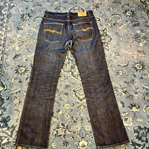 Nudie Jeans Raw Denim - Säljer ett par klassiska mörkblå jeans från Nudie Jeans med snygg färg och tvätt i fint skick med raka ben vilken ger en mer relaxed passform. Tyget är av raw denim vilket gör jeansen tidlösa. Jeansen är i storlek 34/34 men har krympt så dom passar dig som bär ca 32/32. Skriv vid frågor, prioriterar snabb affär så priset går att diskutera.