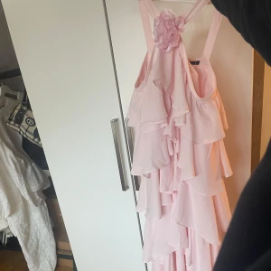 Ljusrosa volangklänning med axelband - Säljer en romantisk ljusrosa långklänning med flera lager volanger och breda axelband. Klänningen har en dekorativ blomma framtill vid axelbanden och ett svepande fall. Perfekt för speciella tillfällen när du vill sticka ut. Aldrig använd, prislapp kvar, passar perfekt till mig som är 169