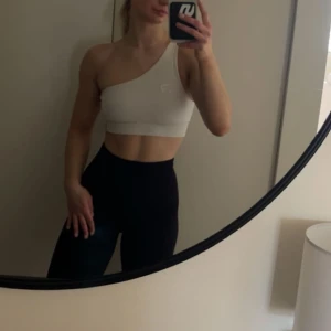 Vit one shoulder träningstopp från Relode - Säljer en vit one shoulder träningstopp från Relode. Toppen är croppad och har en asymmetrisk axelbandslös design, perfekt för gymmet eller andra träningspass. Materialet är stretchigt och bekvämt.