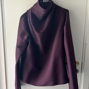 Mörklila topp med draperad krage från Ahlvar Gallery - Säljer en elegant mörklila topp från Ahlvar Gallery med lång ärm och snygg draperad krage. Perfekt för dig som gillar stilrena plagg med en twist. Toppens material känns mjukt och har en fin struktur.