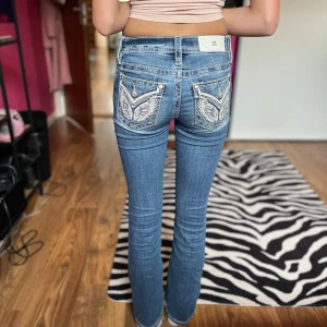 Blå jeansbyxor från Miss Me med broderade detaljer - Säljer ett par blå jeansbyxor från Miss Me, modell med låg midja och snygga broderade detaljer på bakfickorna. Byxorna har bootcut-stil och dekorativa knappar med logga. Perfekta för dig som gillar unika jeans med extra detaljer. Dem är väldigt stretchiga också. Kontakta för mått❤️
