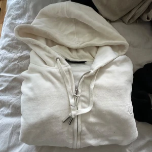Vit hoodie med dragkedja - Säljer en vit hoodie med dragkedja och huva. Tröjan har snörning vid huvan och är tillverkad i mjukt material. Stl M 