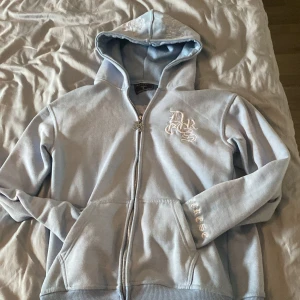 Ljusblå hoodie med dragkedja från Decrese - Säljer en ljusblå hoodie från Decrese med dragkedja och huva. Jackan har broderad logga på bröstet, ärmen och stor brodyr på ryggen. Mjuk bomullskänsla och ribbade muddar vid ärmslut och nederkant.