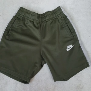 Olivgröna träningsshorts från Nike - Snygga olivgröna shorts från Nike med vita sidor med fina nike detaljer och logga på vänstra ben. Shortsen har elastisk midja och två fickor framtill. Perfekta för träning eller sportiga dagar. Sidorna har en vit rand med Nike-loggor längs hela benet.