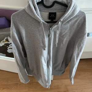 Grå hoodie med dragkedja från Armani Exchange - Säljer en stilren grå hoodie från Armani Exchange med dragkedja framtill och huva med snören. Tröjan har långa ärmar och diskret logga på bröstet. Perfekt för en avslappnad look.