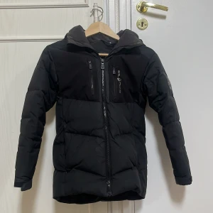 Svart dunjacka från Sail Racing - Säljer en svart dunjacka från Sail Racing med huva och två bröstfickor med dragkedja. Jackan har en stilren design med logga på bröstet och ryggen. Perfekt för kyliga dagar och enkel att matcha med det mesta.