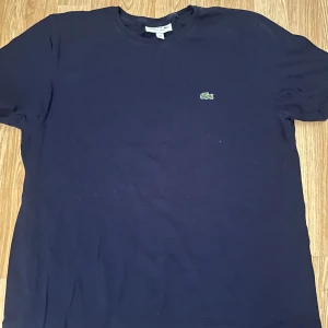 Mörkblå t-shirt från Lacoste - Säljer en mörkblå t-shirt från Lacoste med klassisk rund hals och det ikoniska krokodilmärket broderat på bröstet. Regular fit och tillverkad i mjuk bomull. Perfekt för en stilren och enkel look.